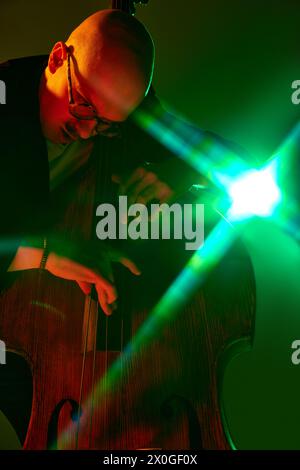Bald talented musician in glasses playing cello in red-green neon against gradient studio background. Stock Photo