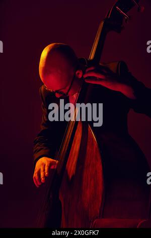Portrait of bald musician in glasses plays double bass in red-yellow light against gradient studio background. Stock Photo