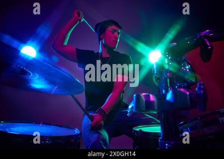 Energetic drummer with drumsticks in pink-purple stage lighting against gradient studio background. Stock Photo