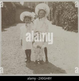 2 girls, about 5 and 8 years old, both wearing white knee-length dresses with black stockings and a large, wide-brimmed white hat with a bow, stand in front of their little brother (about 1 year old), who is dressed all in white and wearing a kind of beret with a black fringed tassel on his head. The children are standing in a driveway to a middle-class house with a flower-covered staircase. They are probably looking at the sun in the shot, as all three of them have their eyes almost closed and the taller girl is grimacing or making a face. [automated translation] Stock Photo