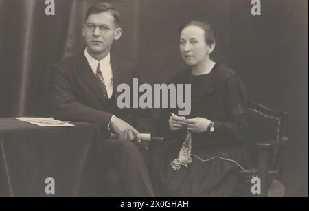 Young man (Arnold costume) approx. 20 years old, side parting, round nickel glasses, wearing a 3-piece black suit, white shirt and narrow striped tie has crossed his knees and is holding a rolled piece of paper in his hand. He rests his arm on a table with a long dark tablecloth. The woman (Anna Födransperg, about 20 years old) is sitting slightly bent forward on a chair, wearing a long black, neckless dress without a corset and holding crocheted items in her hand. She is wearing a watch on her wrist. Her hair is styled back. Both are looking intently past the camera [automated translation] Stock Photo