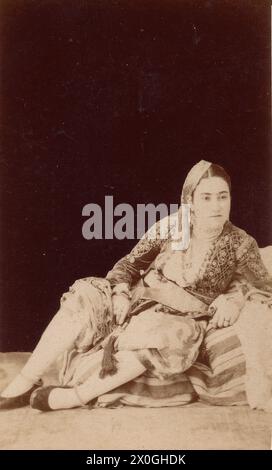 A young oriental-looking woman leans against a bed or high cushion while sitting on the floor. Photograph taken around 1900 at an unknown location. [automated translation] Stock Photo