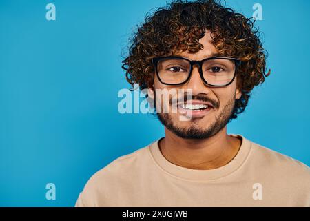 A scholarly Indian student with curly hair and glasses stands confidently against a vibrant blue backdrop. Stock Photo