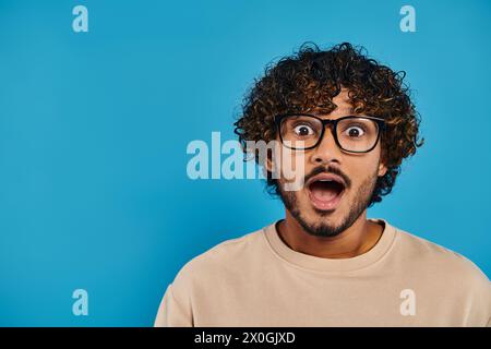 An Indian student with curly hair and glasses looks surprised on a blue backdrop. Stock Photo