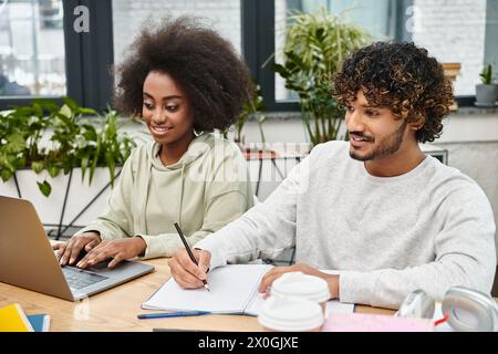 Two people, representing cultural diversity, sit at a table with a laptop, engaged in collaborative study. Stock Photo