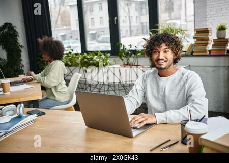 A man of diverse background sits at a table, intensely focused on his laptop in a modern coworking space. Stock Photo