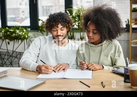 A man and a woman of different ethnic backgrounds sit at a table, writing together in a modern coworking space. Stock Photo