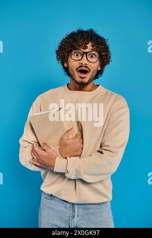 shocked Indian student with curly hair and glasses, standing in casual attire, holding a folder against a blue backdrop in a studio. Stock Photo