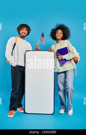 A pair of diverse individuals, multicultural students, stand near a smartphone mockup in a studio against a blue background. Stock Photo