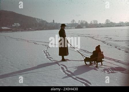 A woman pulls a sledge, on which a small child is sitting, over the frozen Wörthsee. In the background you can see the community of Steinebach and the former lido Fleischmann am Wörthsee. [automated translation] Stock Photo