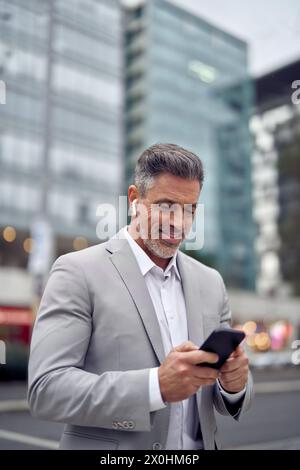 Happy middle aged businessman wearing earbud using phone walking on city street. Stock Photo