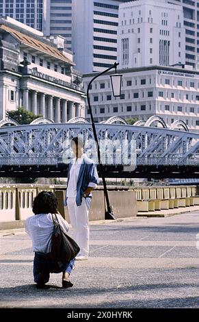 A man poses for a picture on a promenade in front of an impressive scenery in Singapore. He wears white trousers, a white shirt, a blue shirt and puts his hands in his pockets. A woman photographs the scene, kneeling on the ground, wearing jeans, a white blouse and a handbag over her shoulder. In the background there is a white wrought-iron bridge, a streetlamp, on the left a neoclassical building and other modern skyscrapers and skyscrapers. [automated translation] Stock Photo