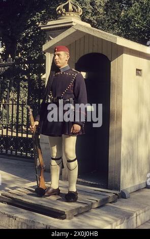 An Evzone in typical uniform stands in front of a guardhouse. Behind it is a fence with a post on which a crown can be seen. [automated translation] Stock Photo
