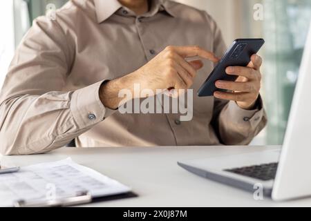 Midsection of a professional man using a smartphone while sitting at a modern office desk with a laptop. Focus on digital communication in a business setting. Stock Photo