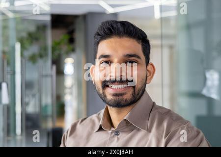 Portrait of a cheerful young man wearing a light brown shirt, smiling confidently in a contemporary office setting. Perfect image for business and lifestyle themes. Stock Photo