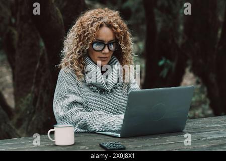 Curly long hair blonde adult woman use laptop computer outdoors sitting in the woods - concept of modern free female peoplelifestyle in smart and remote working activity - online business economy with phone and roaming Stock Photo