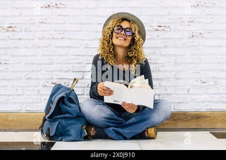 Happy young adult traveler woman sitting on the floor reading a book. Travel female people lifstyle. Curly hair and trendy hat with backpack. Active modern life middle age lady Stock Photo