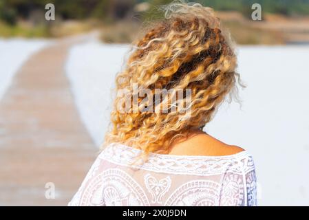 Back view of blonde surf waves hairstyle woman enjoying the beach in summer holiday vacation. Concept of tourist and tourism in the nature. Female people walking on the sand outdoor Stock Photo