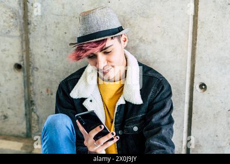 One young modern teenager sitting on the ground using phone app to message and social media life. Online connected youth male boy with trendy fashion alternative clothes in urban life background Stock Photo