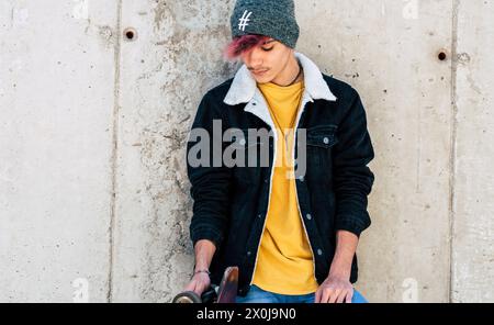 Standing young boy portrait with casual clothes trendy fashion style against a urban wall with skate board. Concept of youth teen lifestyle. Modern generation people posing for picture. Model. Stock Photo