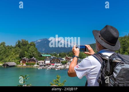 Tourist with black hat and backpack photographed with smartphone, view from Malerwinkel Seterrasse to Königssee, boat landing stage, in the background Unterbergmassiv with the Hochthron, 1972 m, and Almbach Wand, Schönau am Königssee, Berchtesgadener Land, Bavaria, Germany, Europe Stock Photo