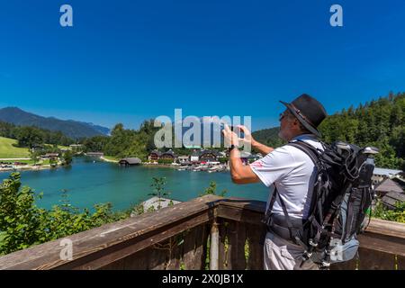 Tourist with black hat and backpack photographed with smartphone, view from Malerwinkel Seterrasse to Königssee, boat landing stage, in the background Unterbergmassiv with the Hochthron, 1972 m, and Almbach Wand, Schönau am Königssee, Berchtesgadener Land, Bavaria, Germany, Europe Stock Photo