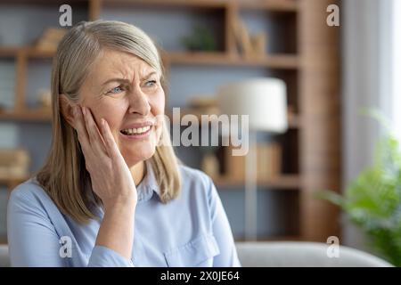 Elderly Caucasian woman suffering from otitis, touching her ear in pain, sitting in a modern living room. She looks distressed and worried about her earache. Stock Photo