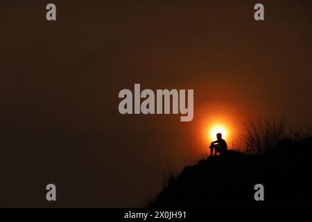 Last sunset of Nepali Year 2080 BS A man sits on the edge of a cliff as the sun sets against the hills surrounding Kathmandu Valley on Nepali new year eve on 12 April, 2024. Nepal on 13 April, 2024 celebrate the new year 2081 BS with grandeur and fervor. Copyright: xSubashxShresthax Stock Photo