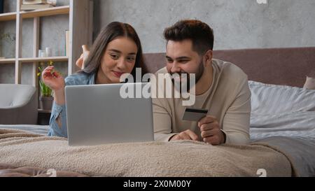 Caucasian happy couple woman and man relax on bed with laptop shopping online use bank credit card pay order buying in internet booking financial Stock Photo