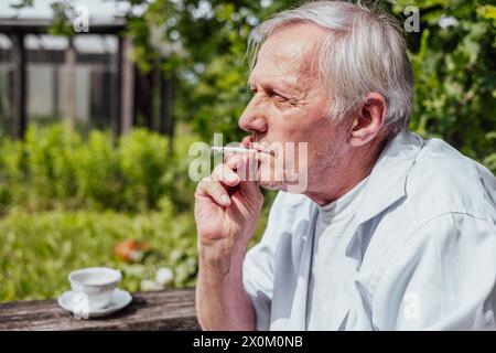 Elderly man smoking a cigarette, wearing a white shirt, outdoors. Reflective, personal moment, suitable for health or lifestyle content. High quality Stock Photo