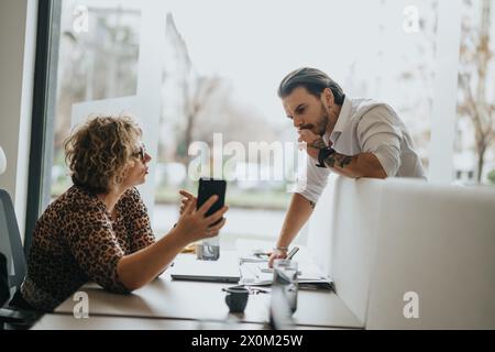 Focused colleagues in a modern office setting engage in a strategic discussion, with one using a smart phone and the other reviewing documents. Stock Photo
