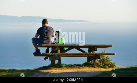 Two hikers rest on a picnic table facing the pacific ocean, enjoy the view. Stock Photo
