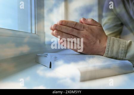 Religion. Double exposure of sky and Christian man praying over Bible at windowsill, closeup Stock Photo