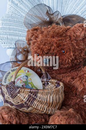 Old teddy bear holds antique egg basket with a 1940s child's record showing children playing. Furry bear sits on an old rattan chair. Display for toys. Stock Photo