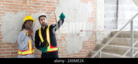 Two construction worker with hardhat and safety vest are examining a wall and ceiling while holding tablet and walkie-talkie. Work atmosphere near sta Stock Photo
