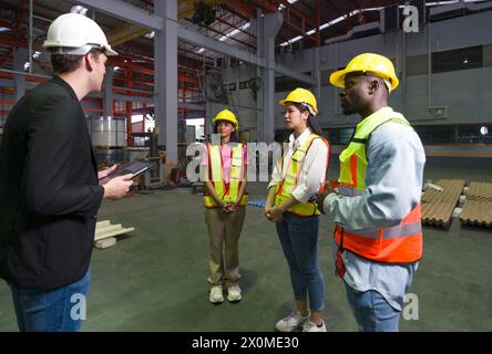 Group of engineer in safety vest meeting with young manager in black suit in an industrial or factory setting. Everyone wearing protective hardhat. At Stock Photo