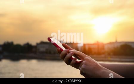 Female person touching screen of her phone with her finger in front of background of city during sunset. Stock Photo