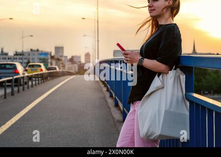 City lifestyle. Urban woman wearing casual outfit stands on the bridge and using her smart phone. Stock Photo