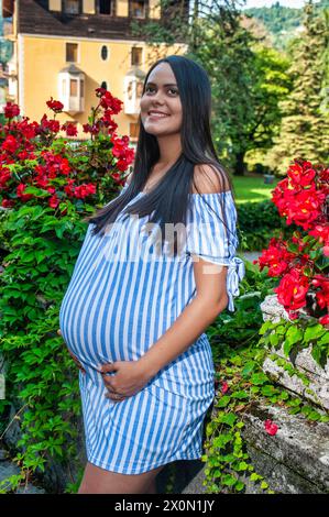 Smiling beautiful pregnant South American girl, amidst flowers, wearing a long striped shirt Stock Photo