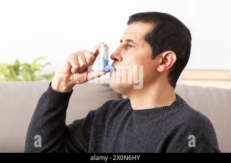 Side view of an asthmatic man using an inhaler sitting on a couch in the living room at home Stock Photo
