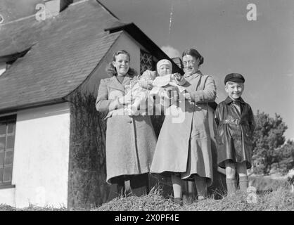 EVACUEES TO A COMMUNAL HOSTEL IN THURLESTONE, SOUTH DEVON, ENGLAND, 1941 - Mrs Findlayson (left, with her 2-month-old daughter Sheila), and Mrs Phillips (with her sons, 9-month-old Roland and 6-year-old Roy) smile for the camera in the sunshine outside the 'Love Nest' in Thurlestone, Devon. Mrs Findlayson and Mrs Phillips have been evacuated to this communal hostel and are caretakers of this rest home, attached to the hostel, which is used by husbands visiting their wives at the Communal Billets whilst home on leave Stock Photo