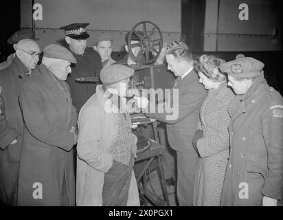 FILM SHOW AT HIGHEST VILLAGE IN THE HIGHLANDS: MINISTRY OF INFORMATION FILM SCREENING, TOMINTOUL, BANFFSHIRE, SCOTLAND, UK, 1943 - Ministry of Information Film Operator John Macdougall explains his Bell & Howell projector to a group of villagers in the Memorial Hall in Tomintoul. Amongst the crowd are the local policeman (third from left) and a member of the local Home Guard (far right). The man in the foreground is smoking a pipe as he inspects the workings of the film equipment Stock Photo