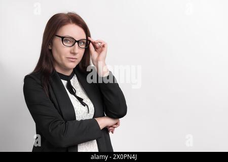 Formal portrait of well dressed businesswoman, wearing jacket and glasses, looking at camera while holding eyepiece of glasses with one hand Stock Photo