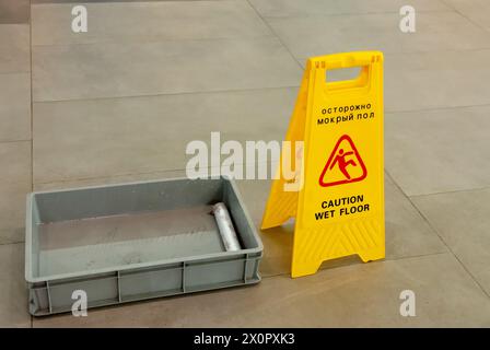 A yellow caution wet floor sign stands beside a gray plastic water tray. A dirty mop in it on a tiled floor, ensuring safety measures are in place dur Stock Photo