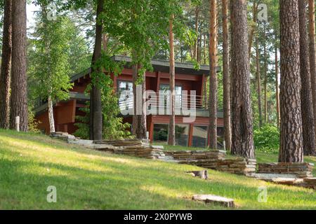 Wooden house in a tranquil pine forest, with balcony, terrace, and lush lawn. Surrounded by tall trees, modern design with large glass windows offerin Stock Photo