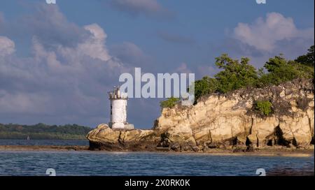 a lighthouse situated on a rocky shore, surrounded by trees, and the scene is set against a backdrop of clouds in the sky with water nearby. Stock Photo