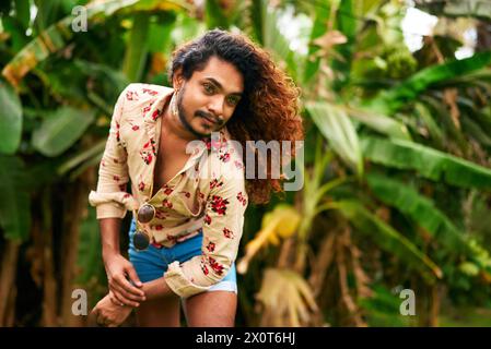 Confident gay man with curly hair in tropical setting poses with style, ethnic LGBTQ individual in floral shirt, denim shorts. Expressive pose in lush Stock Photo