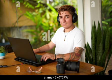 Transgender editor with headphones edits video at laptop in casual office. Focus, determination on face. Camera beside, rich plants background Stock Photo