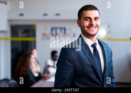 Confident eastern businessman smiles in modern office, diverse team works behind. Arab professional in formal suit leads multicultural group Stock Photo
