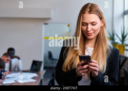 Professional blonde woman in smart casual attire uses smartphone in modern office. Focused businesswoman engages with mobile tech, managing tasks Stock Photo